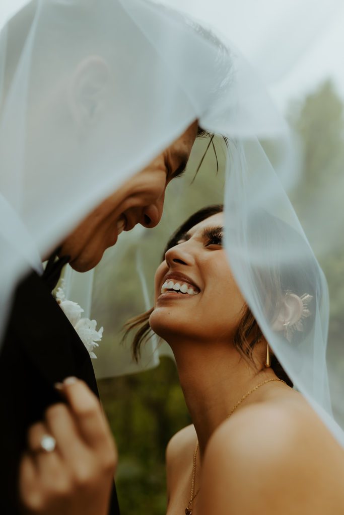 The couple under the veil looking at each other. both have a big smile and bride is gripping the grooms suit jacket collar pulling him in closer to her.