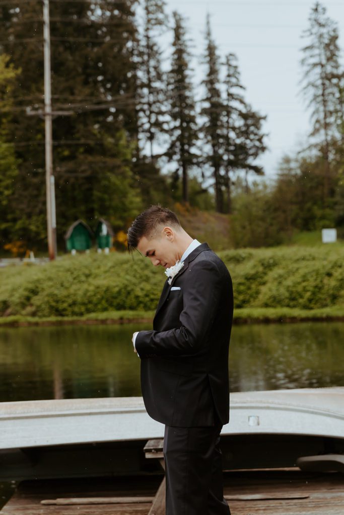 the groom standing on the dock by the pond fixing his suit jacket.