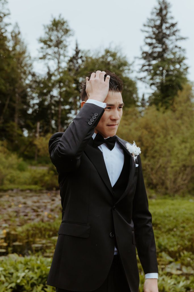 groom standing on the dock by the pond fixing his hair and looking off to the right side