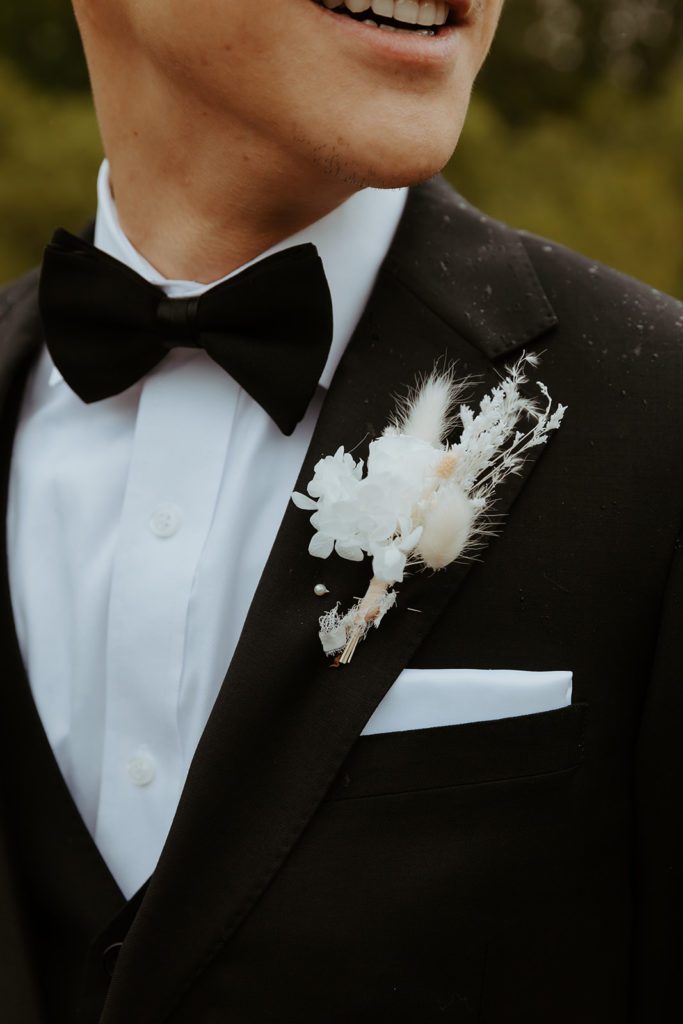 close up of the grooms suit. Wearing a black bowtie has black suit jacket and white dress shirt underneath with a white pocket square. Boutonnière is a mix of white and beige dried florals pinned to the collar of his suit jacket. 