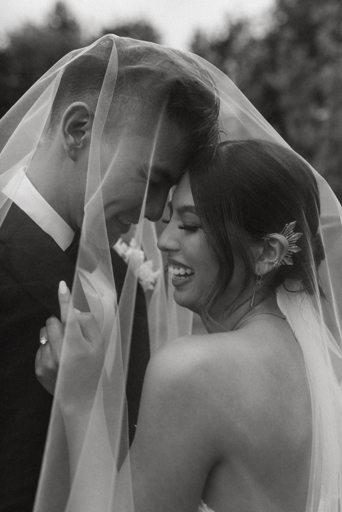 black and white photo of the couple under the veil with foreheads touching. Bride has a big smile and gripping the grooms suit jacket collar pulling him in closer to her.