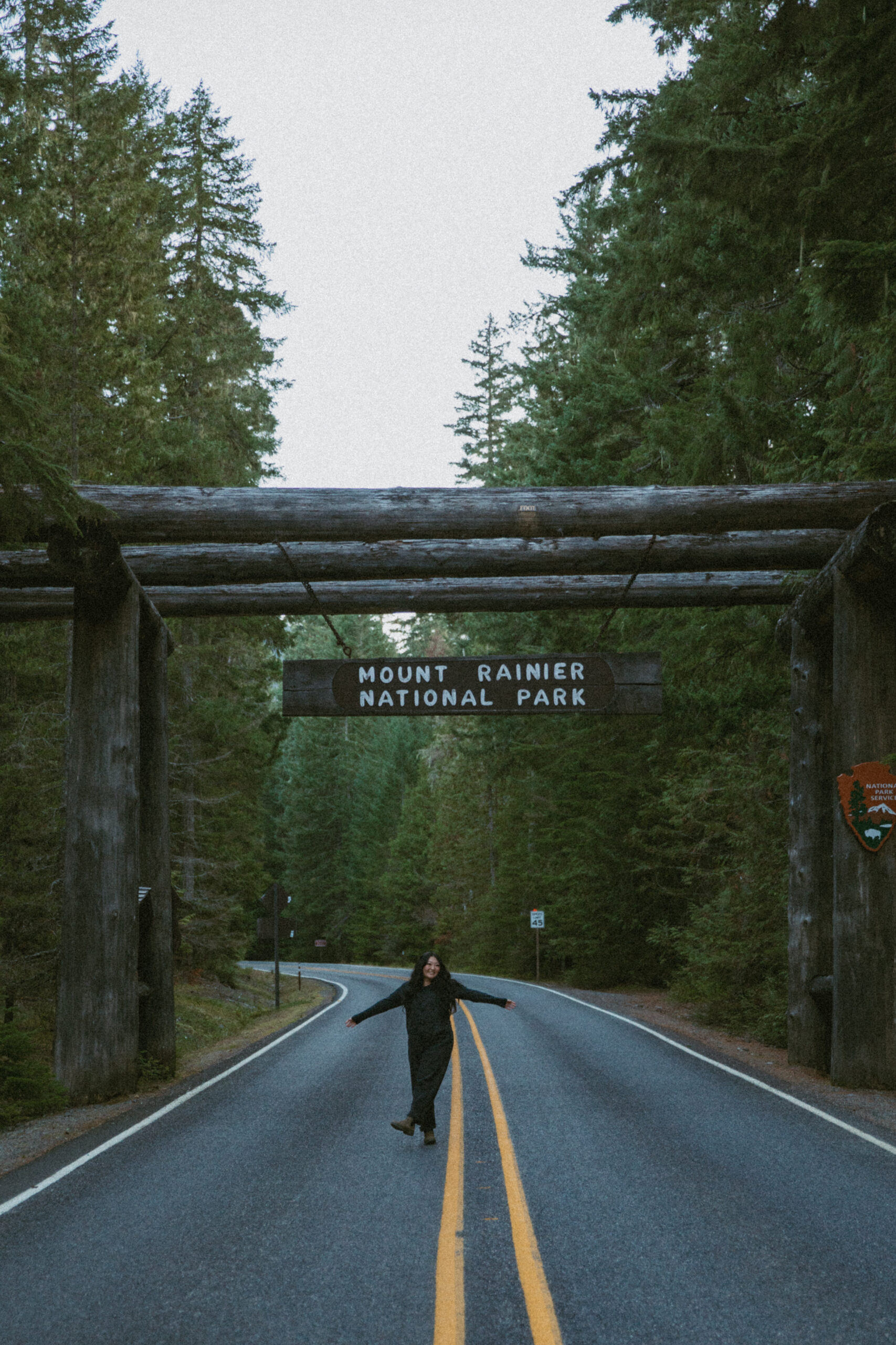girl standing in the middle of the road under the sign at the entrance of mount rainier national park surrounded by tall lush forest pine trees